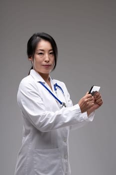 Concerned female Asian doctor or nurse standing holding a mobile phone in her hands as she turns to look at the camera over a grey studio background with copyspace