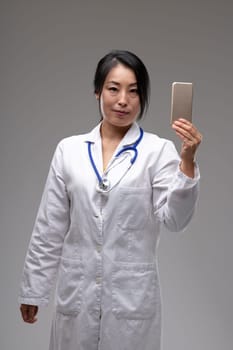 Asian woman doctor in a stethoscope and white lab coat holding up a mobile phone as she looks at the camera over a grey studio background