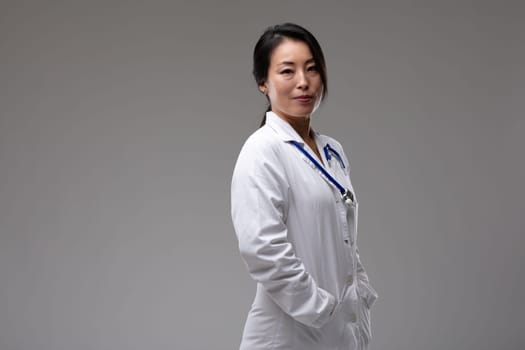 Thoughtful confident Asian woman doctor or nurse in lab coat with stethoscope turning to look at the camera with a quiet smile over grey with copy space