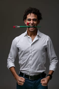 Successful playful confident young artist with daub of red paint on his cheek gripping a paintbrush between his teeth as he clenches his fists in a frontal portrait over a dark studio background