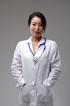 Confident relaxed Asian woman doctor with her hands in pockets in the pockets of her lab coat and stethoscope around her neck isolated on grey