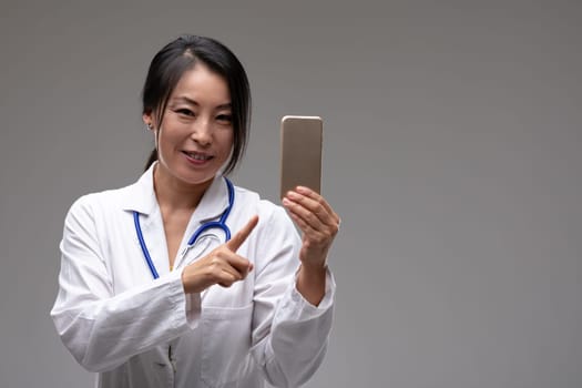 Smiling female Asian doctor or nurse pointing to her mobile phone isolated on grey with copy space