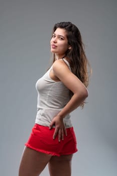 Attractive young woman with long brunette hair wearing bright red sexy skimpy shorts and a sleeveless summer top turning to look back over shoulder at the camera