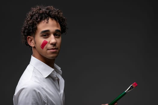 Serious thoughtful young male artist with red paint on cheek turning to look at the camera as he holds a paintbrush in his hand with copyspace over a dark studio background