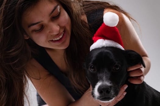 Adorable little dog wearing a red Santa Hat for Christmas and the holiday season posing looking at the camera with his friendly young woman owner
