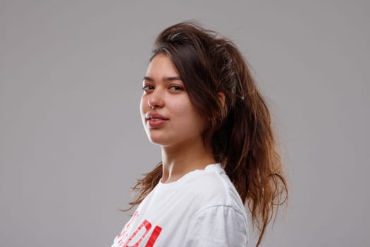Casual young teenage girl with tousled long brown hair posing sideways turning to look thoughtfully at the camera over grey
