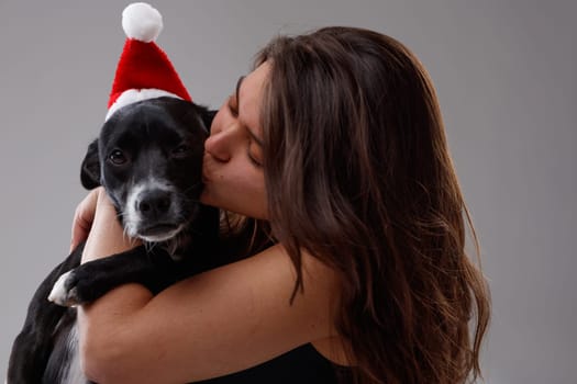 Cute little dog wearing a red Father Christmas hat being kissed by its loving young female owner in a close up portrait celebrating the holiday season