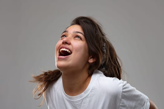 Exuberant young woman vocalising laughing and having fun or screaming in frustration with head tilted back and mouth open over grey