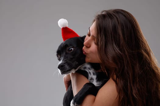 Young woman kissing and hugging a cute little dog wearing a Santa Hat to celebrate the festive season and Christmas