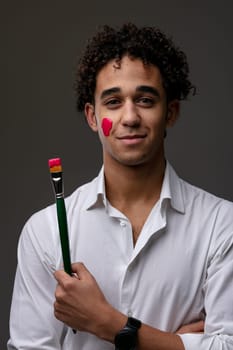 Smiling confident young man with streak of red paint on cheek smiling confidently at the camera while holding a paintbrush in his hand in an art and creativity concept