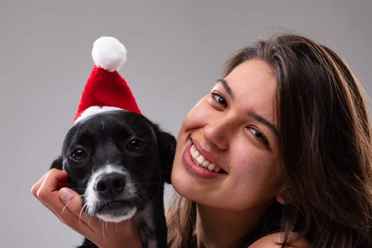 Cute little dog wearing a Santa hat with a smiling young woman alongside to celebrate the Christmas holiday season in a close up on their faces