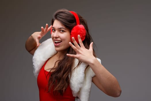 Happy vivacious young woman celebrating the holiday season in colorful festive red ear muffs and matching top with shoulders covered by a stylish white fluffy jacket