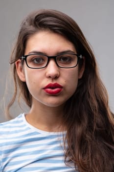 Thoughtful studious young woman wearing glasses staring intently at the camera with parted lips over a grey studio background