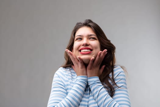 Happy young woman filled with hope and a positive attitude looking up with a joyful smile and hands to her cheeks over a grey studio background