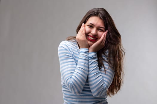 Cute charismatic young woman grinning at the camera with her hands to her cheeks and a beaming smile over a grey studio background