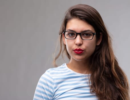 Young dark haired woman wearing glasses staring intently at the camera with a thoughtful expression pouting her red lips over a grey studio background