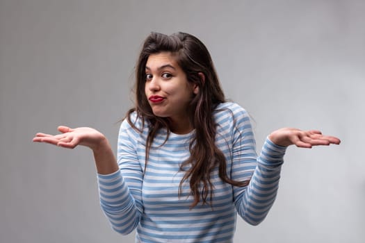 Young woman shrugging her shoulders and gesturing with her hands showing her ignorance or complete disinterest over a grey studio background