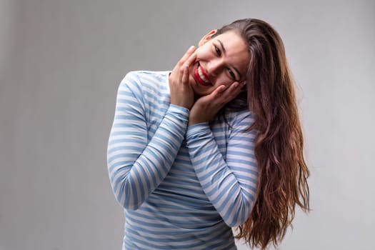 Attractive young woman with a happy sweet smile tilting her head to the side with her hands to her cheeks as she looks at the camera over a grey studio background