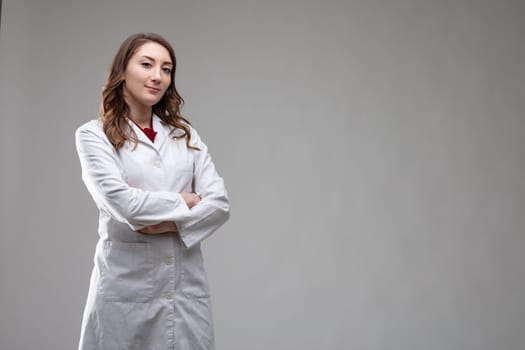 Thoughtful attractive woman doctor in a lab coat with folded arms standing over a grey background looking at the camera with a quiet smile with copy space