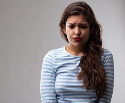Anxious worried moody young woman puckering up her face and frowning as she looks down with a sad expression over a grey studio background