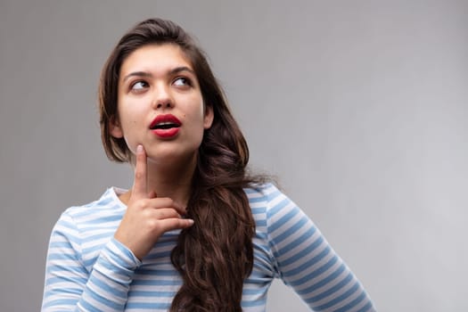 Thoughtful young woman seeking inspiration or trying to solve a problem looking up with a contemplative expression and finger to her chin over a grey studio background