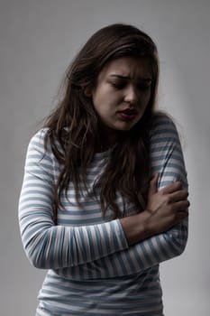 Vulnerable lonely troubled young woman hugging herself with a sad expression and anguished frown over a grey studio background