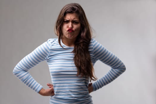 Stubborn young woman puckering up her mouth and glaring at the camera with a frown and hands on hips over a grey studio background