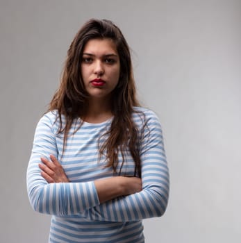Young woman with a deadpan stern expression and penetrating stare standing with folded arms looking at the camera over a grey studio background