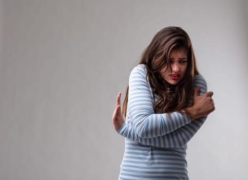 Scared fearful young woman hugging herself protectively and gesturing with her hand to go away or stop cowering to the side over a grey studio background