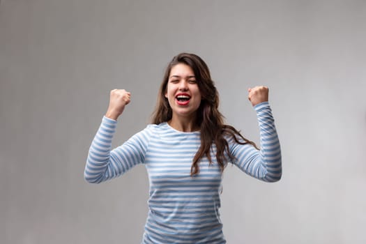 Young woman cheering in exultation after a personal triumph or success punching the air with her fists over a grey background