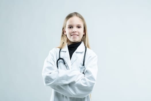 Smiling friendly little girl dressed as a doctor in a white lab coat with stethoscope standing with folded arms grinning at the camera on white with copy space