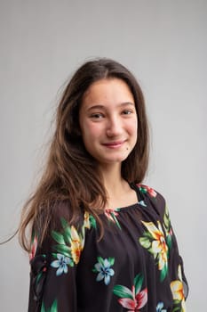 Happy friendly young woman in a floral top turning to smile at the camera with her long hair falling over one shoulder over a grey background