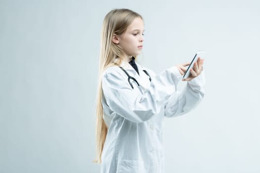 Young girl dressed up in a medical lab coat with stethoscope pretending to be a doctor standing using her mobile phone isolated on white