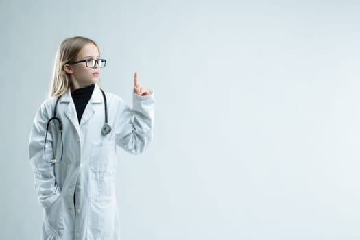 Cute little blond girl playing at being a doctor in lab coat and stethoscope holding up her finger looking towards blank copy space on white