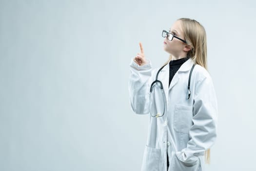 Little girl dressed up as a nurse or doctor in outsize glasses, white coat and stethoscope holding up her finger towards blank copy space on white