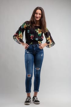 Full length studio portrait of trendy young woman in designer jeans and floral top standing with hands on hips smiling at camera