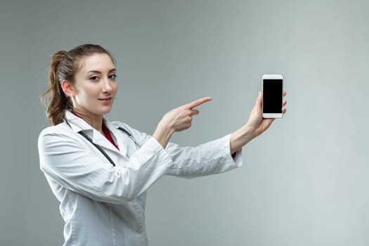 Serious doctor pointing to her mobile phone as she holds it extended in her hand with the blank screen to the viewer over grey with copy space