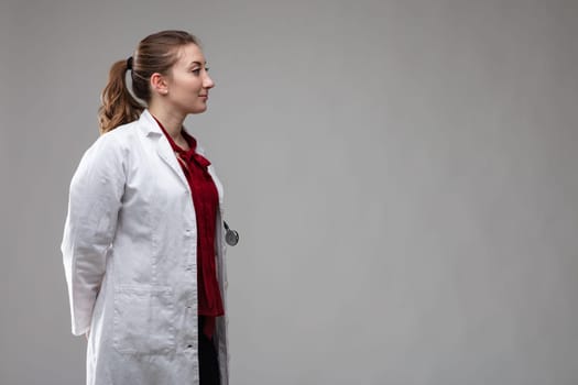 Profile portrait of a woman doctor or nurse in a white lab coat and stethoscope looking towards blank copyspace on grey