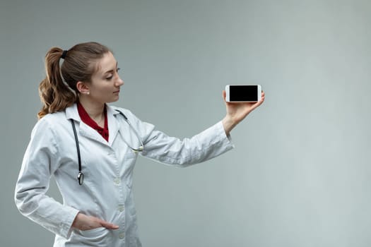 Woman doctor or nurse wearing a stethoscope and white lab coat standing holding up her mobile phone with the screen to the camera over grey wth copy space