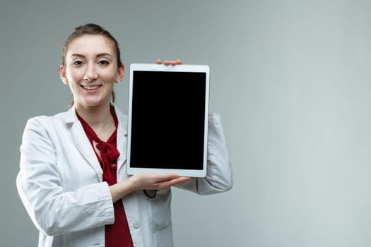 Happy smiling woman doctor holding up a blank tablet pc with the screen to the viewer over a grey studio background with copy space