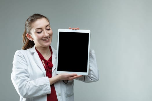 Smiling friendly woman doctor or nurse holding up a tablet on view to the viewer over a grey background with copy space