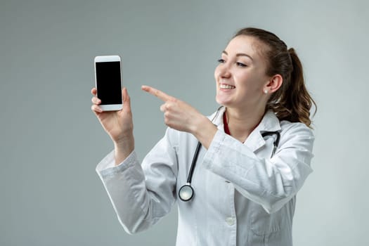 Woman doctor or nurse wearing a lab coat and stethoscope pointing to a blank mobile phone with a pleased smile close up over a grey background