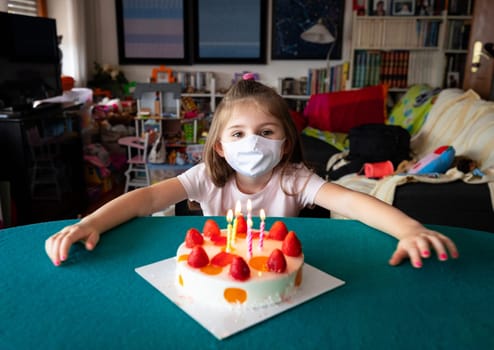 Little five year old girl wearing a protective face mask during the coronavirus or Covid-19 pandemic seated at table with arms outstretched around her birthday cake with burning candles