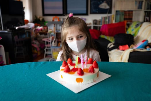 Pensive little five year old girl celebrating her birthday seated in a protective face mask at table looking quietly at her cake topped with strawberries and unlit candles during the Covid-19 pandemic