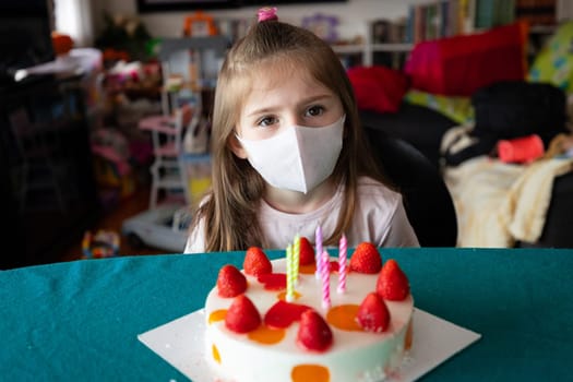 Young girl celebrating her birthday in a face mask during the coronavirus or Covid-19 pandemic seated at a table in front of a strawberry cake with candles
