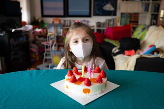Thoughtful little birthday girl wearing a protective face mask during the coronavirus or Covid-19 pandemic sitting at table celebrating at home with a delicious fruity cake with candles