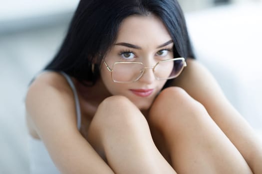 Soft focus dreamy portrait of a beautiful young woman wearing trendy glasses sitting clasping her legs looking up at the camera with wide brown eyes and a calm expression in a cropped portrait