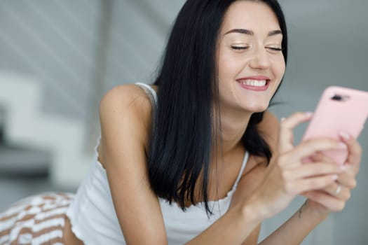 Happy young woman with a joyful vivacious grin using her mobile phone as she relaxes at home in a close up view on her face
