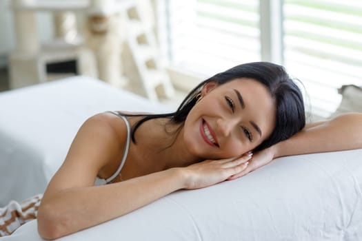 Beautiful young woman with a lovely sincere friendly smile relaxing resting her head on the arm of a sofa smiling happily at the camera in a high key portrait