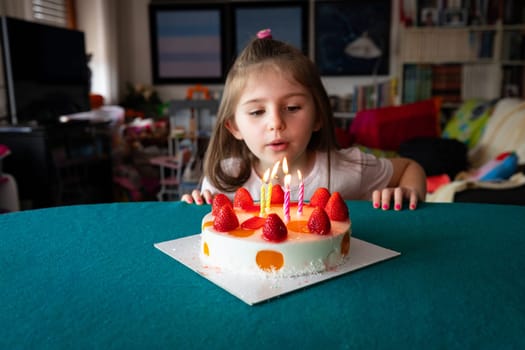Adorable little girl blowing out five candles on her birthday cake topped with fresh ripe strawberries at home in the living room with copy space in a close up portrait